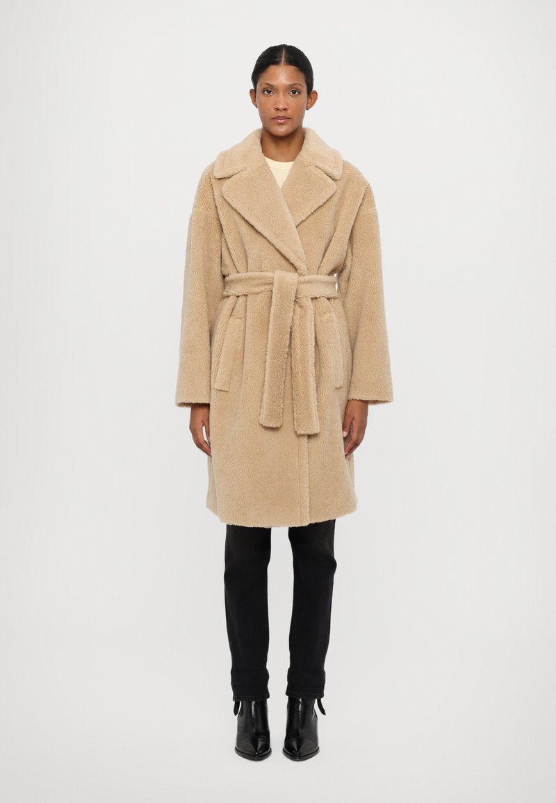 Beige, belted, knee-length coat made of plush, textured fabric. Features wide lapels and long sleeves, paired with black fitted pants and heeled boots.