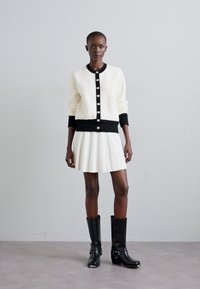 Gilet blanc avec des bordures noires et de gros boutons, associé à une jupe blanche plissée et des bottes noires jusqu'aux genoux avec du matériel décoratif.