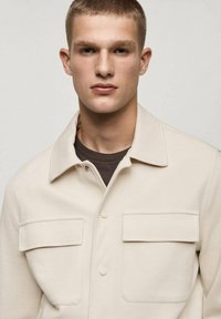 Light beige button-up shirt made of textured fabric, featuring a collar, two chest pockets, and long sleeves with a brown undershirt underneath.