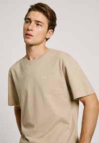 Beige cotton t-shirt with a round neckline, short sleeves, and a small white logo on the left chest. Soft texture, casual fit.