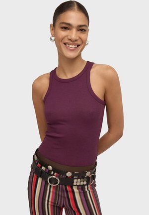 Nasmejana žena s kosom kose vezanim unazad nosi ljubičastu bluzu bez rukava i prugaste hlače s crnim pojasom sadržeći srebrne zakovice i prstenove.