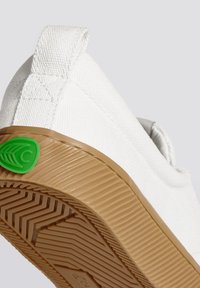 Scarpa da ginnastica bianca in tela con una finitura testurizzata, suola in gomma color biscotto e un'accensione del logo verde sul tallone. Presenta un'etichetta sul tallone per un facile indosso.