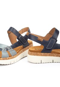 Las sandalias con suela de corcho presentan una parte superior de cuero azul claro con un diseño entrecruzado y correas azul oscuro. Suela de goma blanca con acabado texturizado.