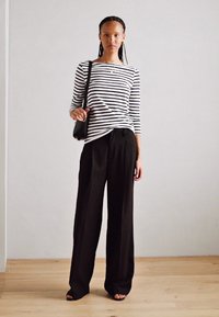 Gestreiftes schwarz-weißes Langarmshirt, kombiniert mit hoch taillierten, schwarzen, weiten Hose, Model hält eine kleine schwarze Handtasche.