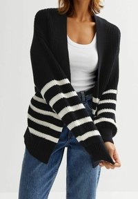 Svart stickad kofta med vita horisontella ränder, öppen fram, och långa ärmar. Har ribbade muddar och en lös, bekväm passform.