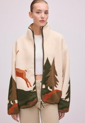 Femme portant une veste polaire beige avec un motif forêt et cerfs vert et marron, zippée à moitié, associée à un crop top blanc et un pantalon beige.
