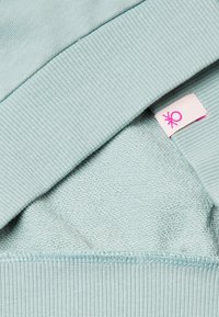 Svetle modrá mikina s vroubkovaným límcem a spodním lemem, s měkkým, texturovaným interiérem. Obsahuje růžovou náušnici s ženským symbolem.