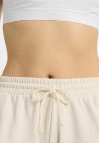 Pantalon de survêtement couleur crème avec une taille élastique et un cordon de serrage, associé à un crop top blanc ajusté. Texture lisse visible.