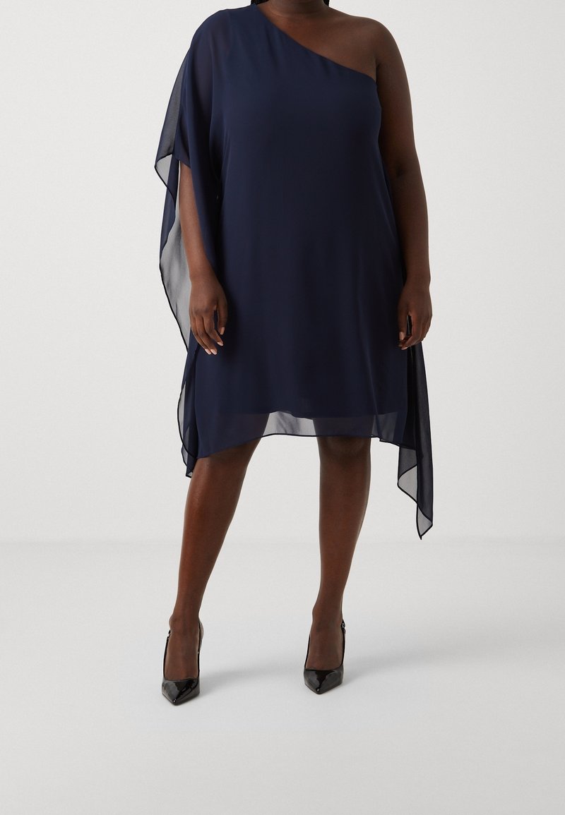 Femme grande taille portant une robe marine transparente à une épaule avec un ourlet asymétrique et des escarpins noirs à bout pointu, debout sur un sol blanc.