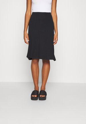 Femme portant une jupe noire longueur genoux, un haut blanc sans manches et des sandales compensées noires sur un fond clair uni.
