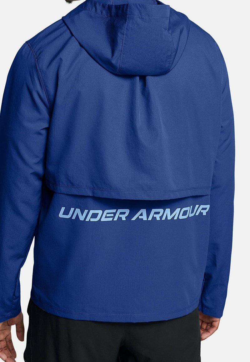 Chaqueta con capucha azul hecha de material ligero. Presenta un gran logo de "UNDER ARMOUR" en la parte posterior en azul claro. Diseño limpio con textura suave.