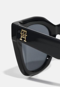Ochelari de soare negri cu un cadru geometric, lentile întunecate și un accent cu logo auriu pe lateral. Material plastic neted și lucios.