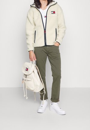 Crème fleece zip-up jack met donkere afwerking, olijfgroene broek, witte sneakers en een lichtbeige drawstring rugzak met een logo patch.