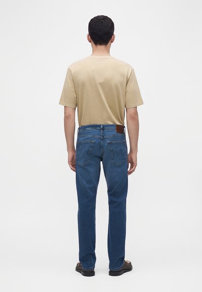 Homme debout de dos, portant un t-shirt beige, un jean bleu avec des poches arrière et des chaussures foncées sur un fond uni.