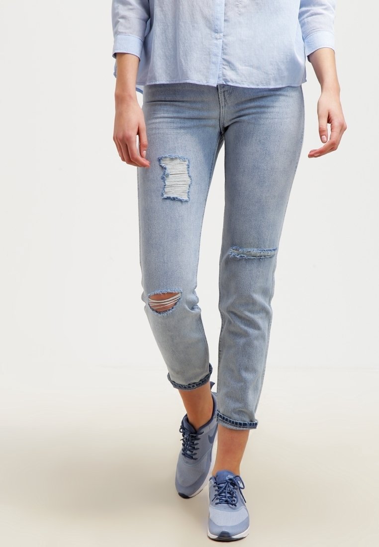 Henkilö kävelee yllään vaaleansiniset repaleiset farkut, vaaleansininen napitettava paita ja siniset nauhalla varustetut lenkkarit.