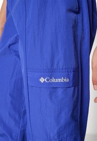Bolso azul da Columbia com um emblema visível, usado por uma pessoa com a mão parcialmente visível do lado direito.