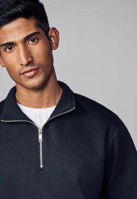 Zwart half-zip pullover met een gestructureerd oppervlakte, contrasterende witte binnenlaag en zilveren ritsdetails. Vloeiende kraag en mouwen.