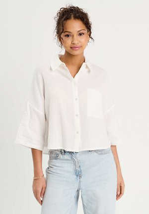 Jonge vrouw met krullend haar, gekleed in een losse witte blouse en lichtblauwe hoge taille spijkerbroek, staand tegen een effen achtergrond.