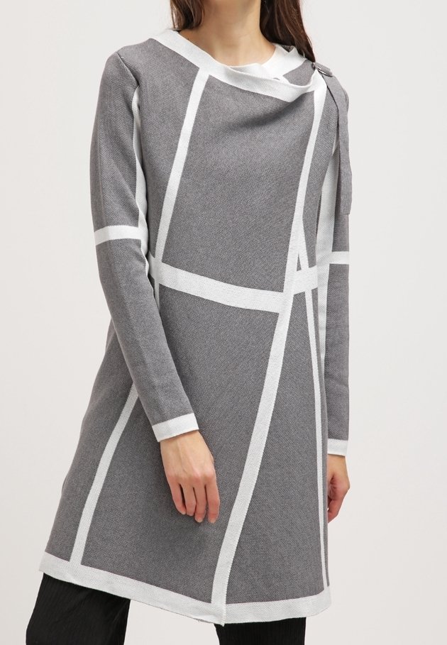 Femme portant une robe grise à manches longues avec des lignes géométriques blanches et un décolleté asymétrique, associée à un pantalon noir.