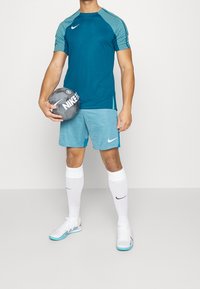 Man i teal sportskläder som håller en grå Nike fotboll, klädd i ljusblå shorts, vita strumpor och vita inomhusfotbollsskor.