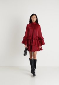 Rød gjennomsiktig kjole med volanger, høy hals og lange ermer. Kombinert med glansfulle, svarte støvler til knærne og en svart naglet håndveske.