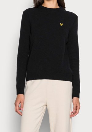Femme portant un pull noir à manches longues avec un petit logo d’oiseau jaune sur la poitrine, associé à un pantalon beige clair.