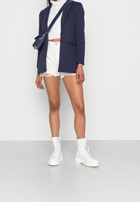 Marinblå blazer, vit kortstickad tröja, slitna vita shorts och vita snörade stövlar i knähöjd med kraftiga sulor. Axelremsväska i blått.