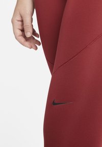 Czerwone legginsy sportowe o gładkiej fakturze, z czarnymi akcentami logo Nike i detalami szwów, które zapewniają dopasowany krój.