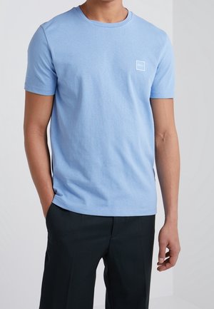 T-shirt en coton bleu clair avec un col rond, des manches courtes et un petit badge logo blanc sur la poitrine, présenté avec un pantalon sombre.