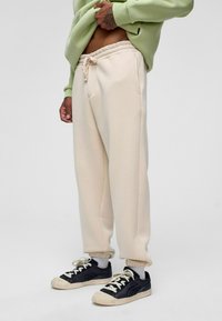 Beige sweatpants met taps toelopende enkels en een verstelbare tailleband met koord, gepaard met zwarte sneakers met witte accenten en veters.