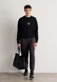 Modelis melnā sporta džemperī ar kvadrātveida emblēmu, pelēkās sporta biksēs ar sarkano logo un melnā auduma somā. Apavi - melni sneakers.