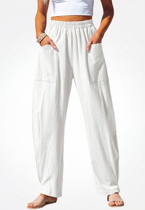 Vrouw draagt losse witte broek met grote voorzakken, een wit cropped top, armbanden en beige open-toe sandalen, staand met handen in de zakken.