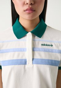 Polo com colarinho verde, corpo creme e riscas horizontais azul claro. Logótipo da Adidas em verde no lado esquerdo. Tecido texturizado.