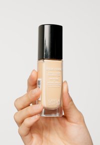 Fondotinta in una bottiglia di vetro trasparente con un tappo nero. Liquido beige chiaro visibile. L'etichetta riporta la dicitura "HYDRO TENSE" e include i dettagli del prodotto.