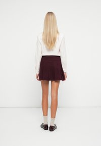 Vrouw met lang blond haar die een witte blouse met lange mouwen, een donkerpaarse geplooide rok, beige sokken en donkerrode schoenen draagt, staand met haar rug naar de kijker gekeerd.