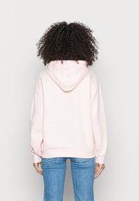 Persona con capelli ricci che indossa un hoodie rosa chiaro e jeans blu, in piedi rivolto verso l'esterno contro uno sfondo chiaro e uniforme.