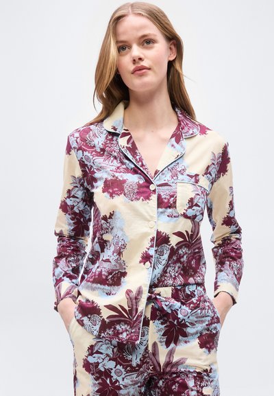 Γυναίκα με πουκάμισο μακρυμάνικο με κουμπιά σε κρεμ, μπορντό και ανοιχτό μπλε, με λουλουδάτο μοτίβο, και αντίστοιχο παντελόνι, με τα χέρια στις τσέπες, ουδέτερο φόντο.