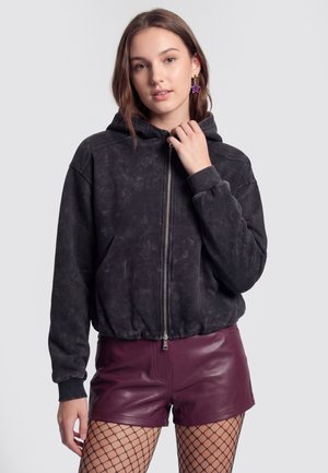 Junge Frau trägt einen schwarzen Zip-Hoodie, burgunderrote Leder-Shorts, Netzstrumpfhosen und lila sternförmige Ohrringe.