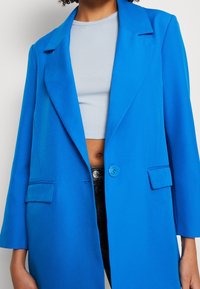 Femme portant un blazer bleu vif à un seul bouton par-dessus un crop top gris clair et un pantalon noir, debout devant un fond blanc uni.