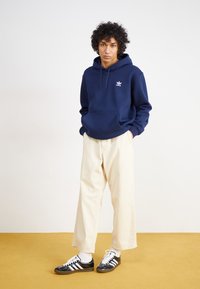 Marineblauwe hoodie met voorzak en wit logo, gecombineerd met beige wijde broek en zwarte sneakers met witte strepen.