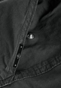 Cămașă din țesătură gri, cu o cusătură vizibilă, capse metalice și un eticheta logo de culoare neagră cu designuri geometrice. Textură netedă cu un finisaj mat.