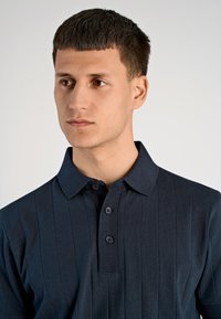 Granatowa, prążkowana koszulka polo z kołnierzykiem, trzema guzikami i teksturowanym materiałem. Zbliżenie na koszulkę na górnej części ciała modela.