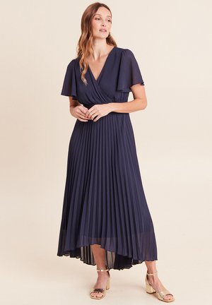 Frau trägt ein marineblaues plissiertes Midikleid mit ausgestellten Ärmeln und goldenen Blockabsatz-Sandalen mit Knöchelriemen, steht vor einem schlichten Hintergrund.