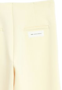 Pantalones color crema con una textura suave, dos bolsillos traseros y una etiqueta cosida debajo del bolsillo derecho. Sin cierres visibles.