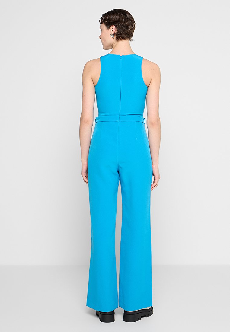 Vrouw met kort haar in een mouwloze felblauwe jumpsuit en zwarte plateau-schoenen, staand met de rug naar de camera tegen een effen achtergrond.