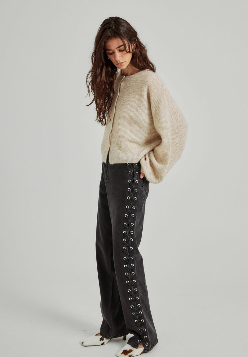 Cárdigan beige de punto con cuello redondo, mangas oversize, combinado con pantalones negros que presentan cordones laterales y ojetes plateados.