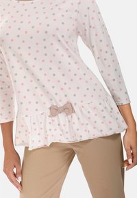Weißes Langarmoberteil mit rosa und grauen Punkten, rüschensaum und einem kleinen Schleifenakzent. Kombiniert mit beigen, engen Hosen.
