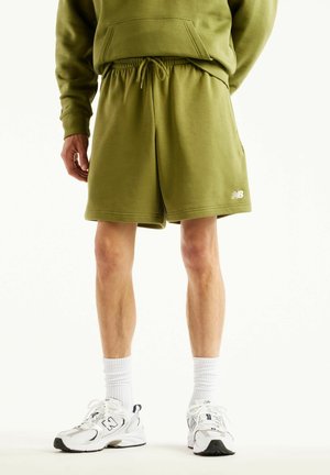 Uomo che indossa pantaloncini verde oliva con coulisse, calzini bianchi alla caviglia e sneakers bianche New Balance, in piedi su sfondo bianco.