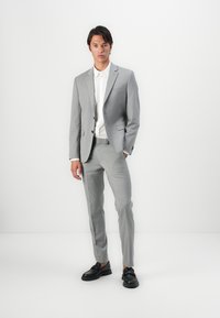 Costume ajusté gris clair avec un motif texturé subtil, associé à une chemise blanche à col et des chaussures noires. Coupe moderne et silhouette slim.