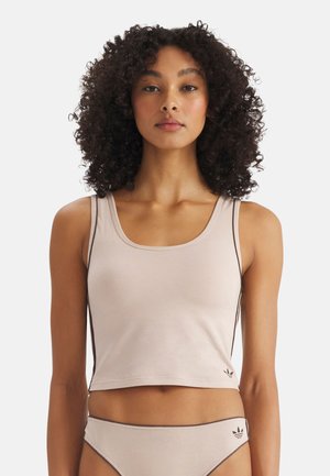 Vrouw met krullend haar die een lichtbeige cropped tanktop en bijpassende slip met donkere rand en klein Adidas-logo draagt.
